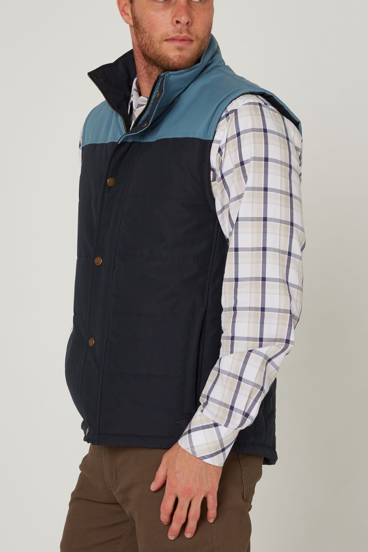 A man in a blue and black Pilbara Two Tone Vest with polyfill padding over a white plaid shirt and brown pants stands sideways, looking slightly left, his left hand resting by his side.