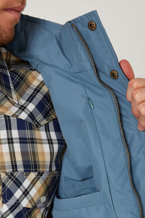 A person models the Pilbara Two Tone Vest, holding it open to display its inner lining and pocket details, worn over a blue, beige, and white plaid shirt.
