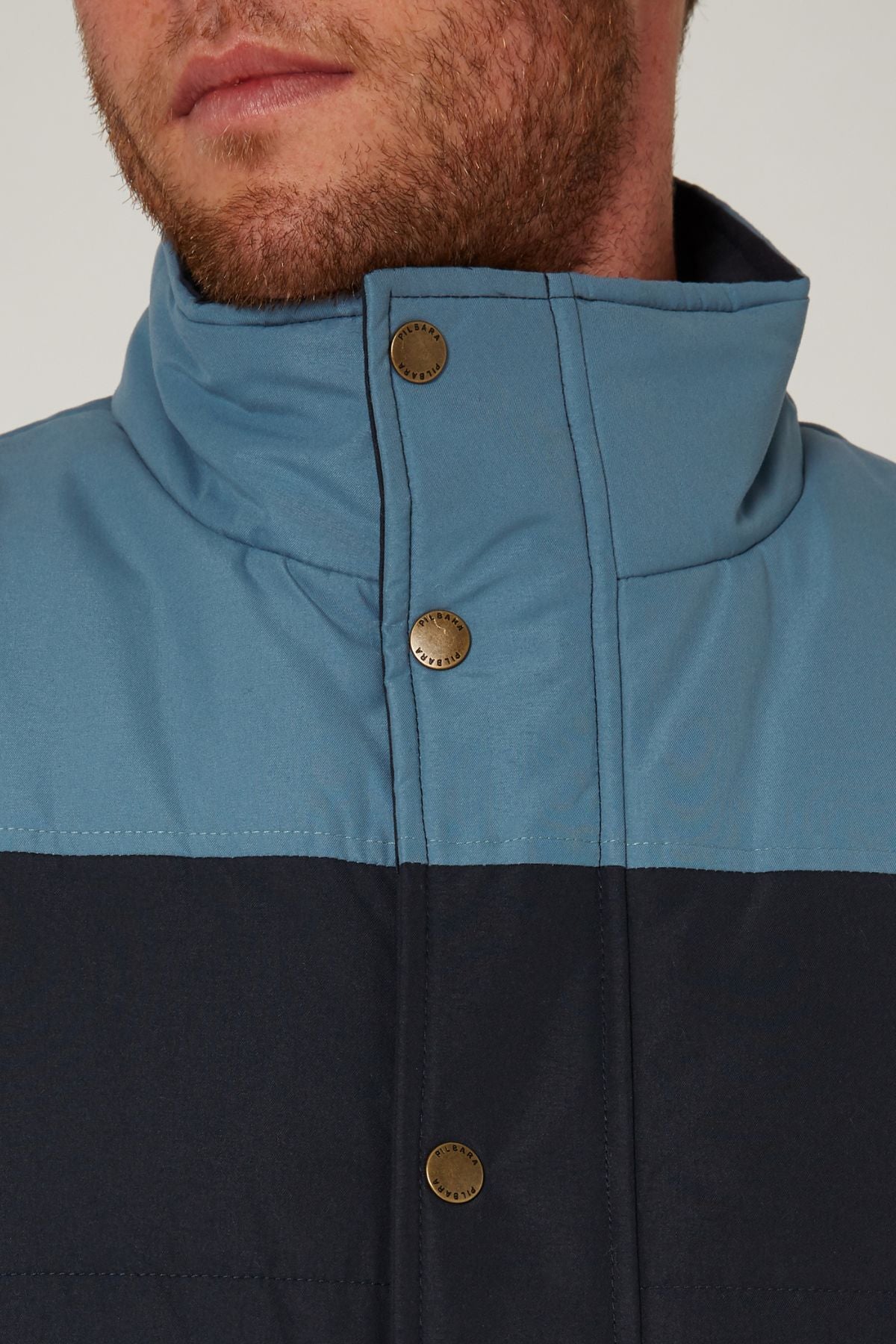 A close-up of a man's lower face and neck shows the Pilbara Two Tone Vest in blue and black, featuring metal snap buttons, polyfill padding, and a high collar. The man has light skin and a short beard.