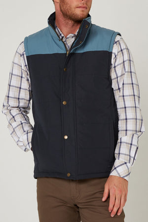 A man wears the Pilbara Two Tone Vest in blue and navy with polyfill padding over a plaid button-down shirt and brown pants, standing with one hand in his pocket.