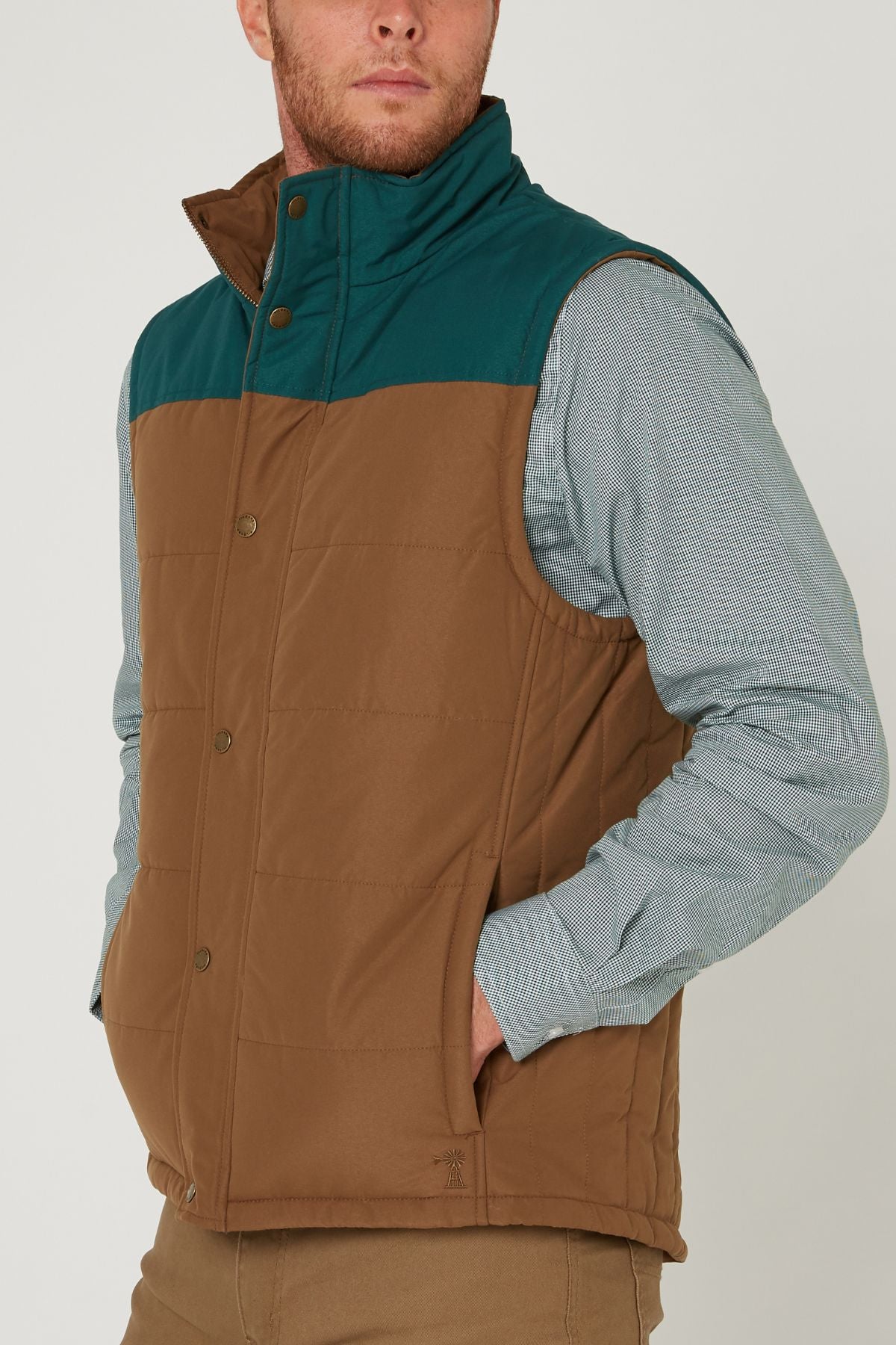 A man models the Pilbara Two Tone Vest in brown and teal over a light blue, long-sleeve shirt and tan pants, standing with his hand in his pocket and looking slightly to the side.