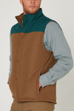 A man models the Pilbara Two Tone Vest in brown and teal over a light blue, long-sleeve shirt and tan pants, standing with his hand in his pocket and looking slightly to the side.