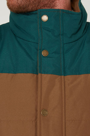 Close-up of a person wearing the Pilbara Two Tone Vest, featuring a teal upper section, brown lower section, high collar, and metal snap buttons. Only part of the lower face and neck is visible.
