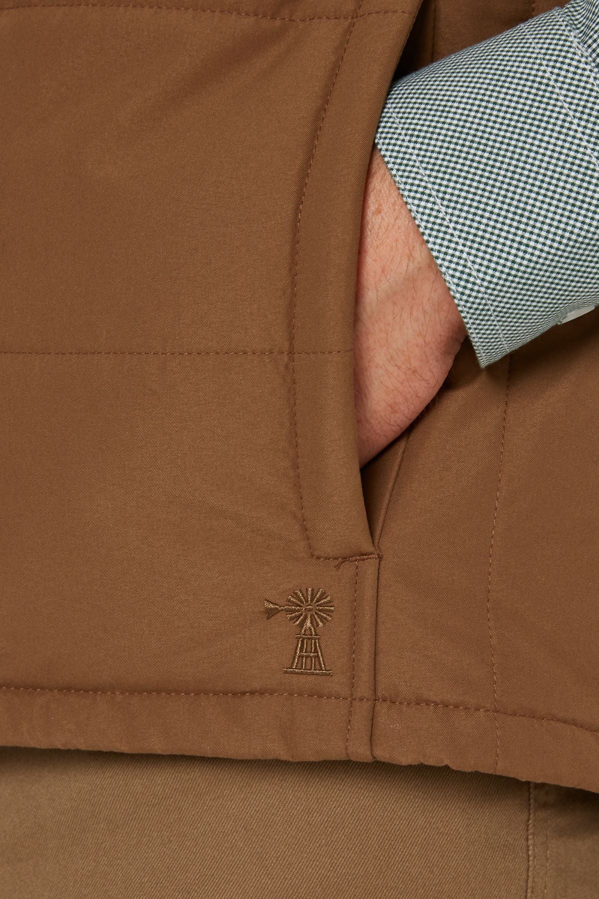 A close-up of a hand in the pocket of the Pilbara Two Tone Vest, featuring a small windmill embroidery. The person pairs it with a blue and white checkered shirt and brown pants.