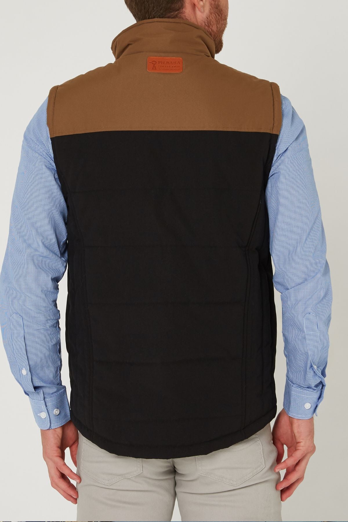 A man is seen from behind wearing the Pilbara Two Tone Vest, a classic fit padded brown and black vest with polyfill fill, over a light blue button-up shirt and beige pants. The vest features a brown patch on the upper back near the collar.