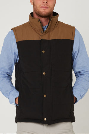 A man wears the Pilbara Two Tone Vest—a brown and black classic fit quilted vest with polyfill padding, stand-up collar, gold snap buttons, and pockets—over a light blue button-down shirt.