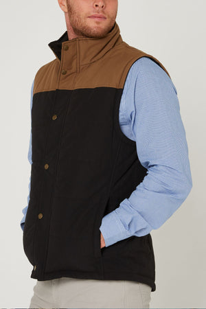 A man stands against a plain background wearing a light blue button-up shirt, beige pants, and the Pilbara Two Tone Vest—a classic fit waterproof vest with a brown top section and black lower section, one hand in his pocket.