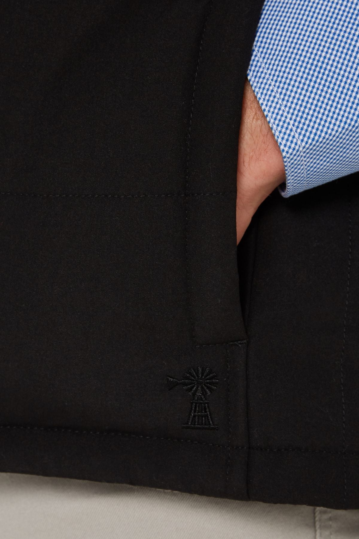 A person wearing a blue checkered shirt and light pants has their hand in the pocket of the Pilbara Two Tone Vest, a classic fit black vest featuring a small embroidered windmill design near the pocket.