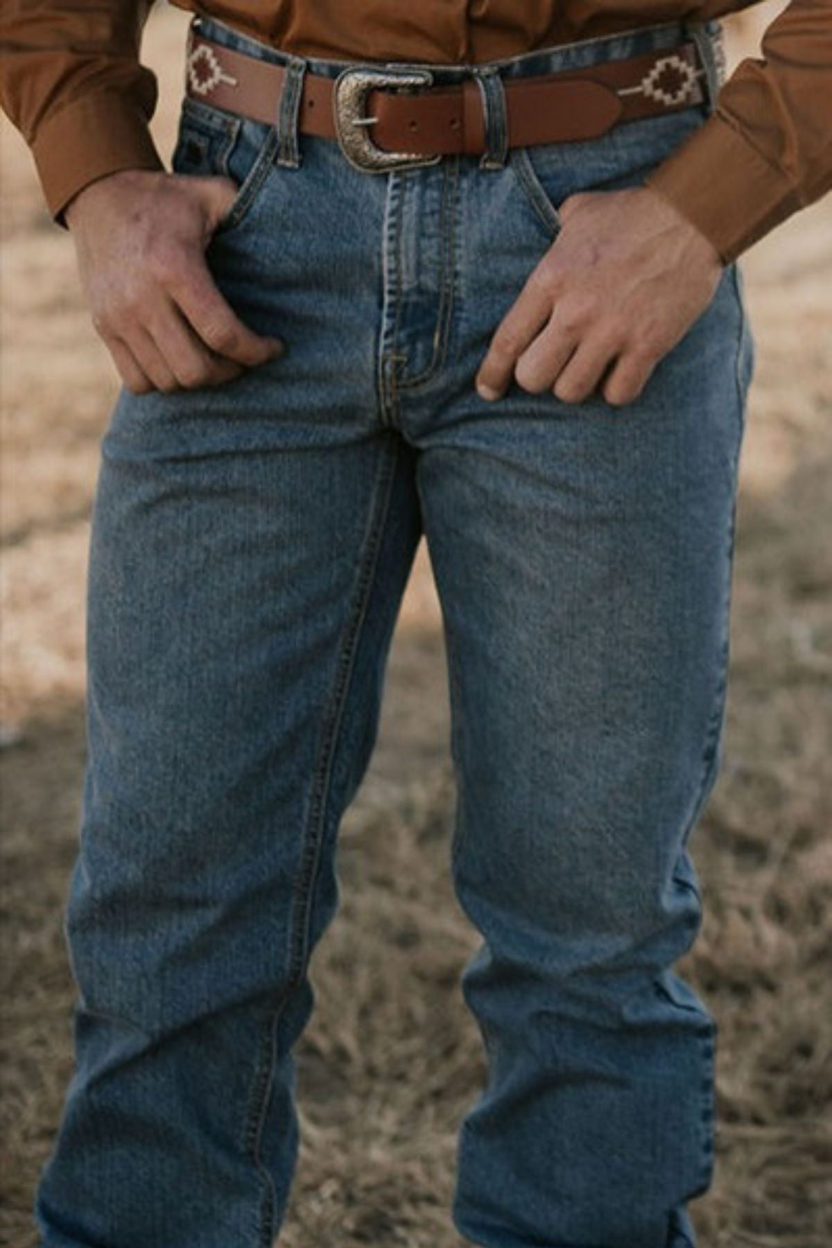 Wearing Pilbara Western Angus Jeans in indigo wash, complemented by a brown leather belt featuring a decorative pattern, and accompanied by a premium quality brown shirt, the person stands outdoors on the grass with hands confidently resting on their hips.