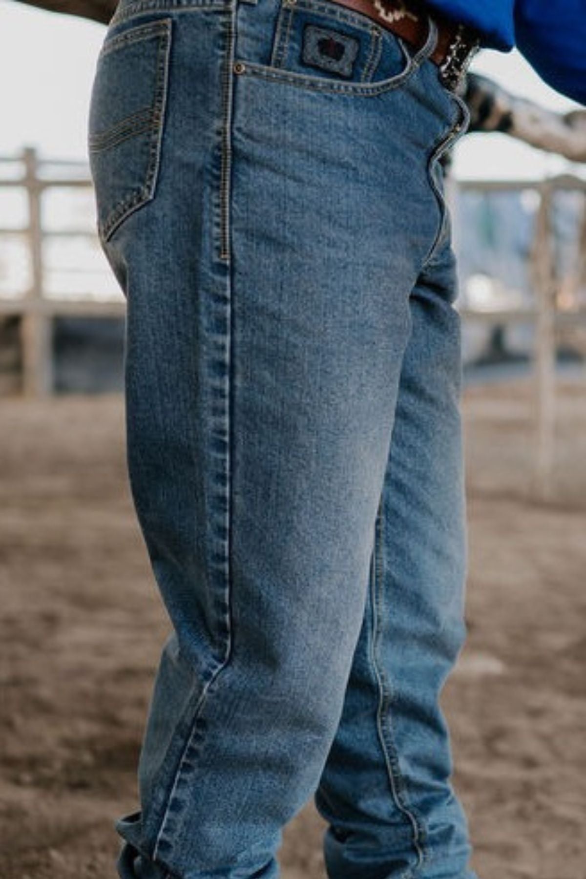 A person wearing the Pilbara Western Angus Jeans stands in an outdoor setting. The background reveals a rustic farm area with wooden fences, while the focus remains on the jeans, emphasizing their indigo wash and impeccable fit.