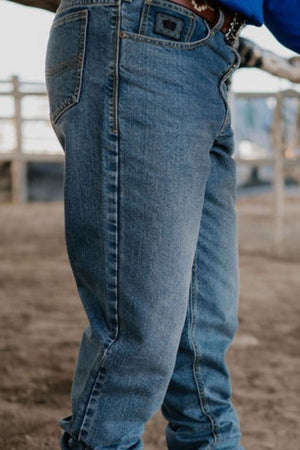A person wearing the Pilbara Western Angus Jeans stands in an outdoor setting. The background reveals a rustic farm area with wooden fences, while the focus remains on the jeans, emphasizing their indigo wash and impeccable fit.