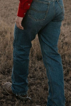 A person wearing Pilbara Western Angus Jeans in premium quality indigo wash, along with a red shirt and cowboy boots, stands on dry grass. Only their lower body is visible, with one foot slightly raised.