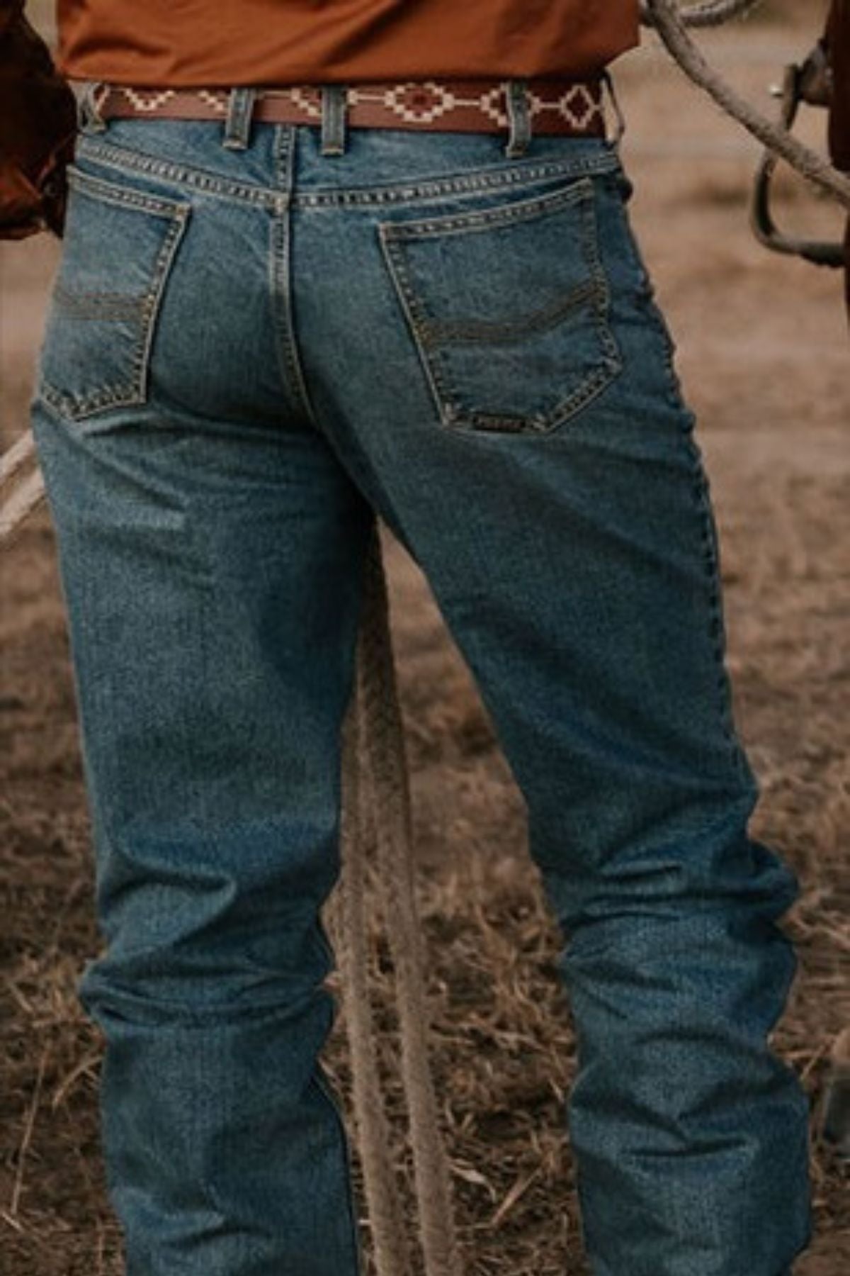 A person wearing Pilbara Western Angus Jeans, known for their premium quality and indigo wash, stands on dry grass while holding a rope. They are accessorized with a brown belt featuring a decorative pattern. The jeans include pockets and have a leather patch on the back as the scene unfolds outdoors in a rural setting.