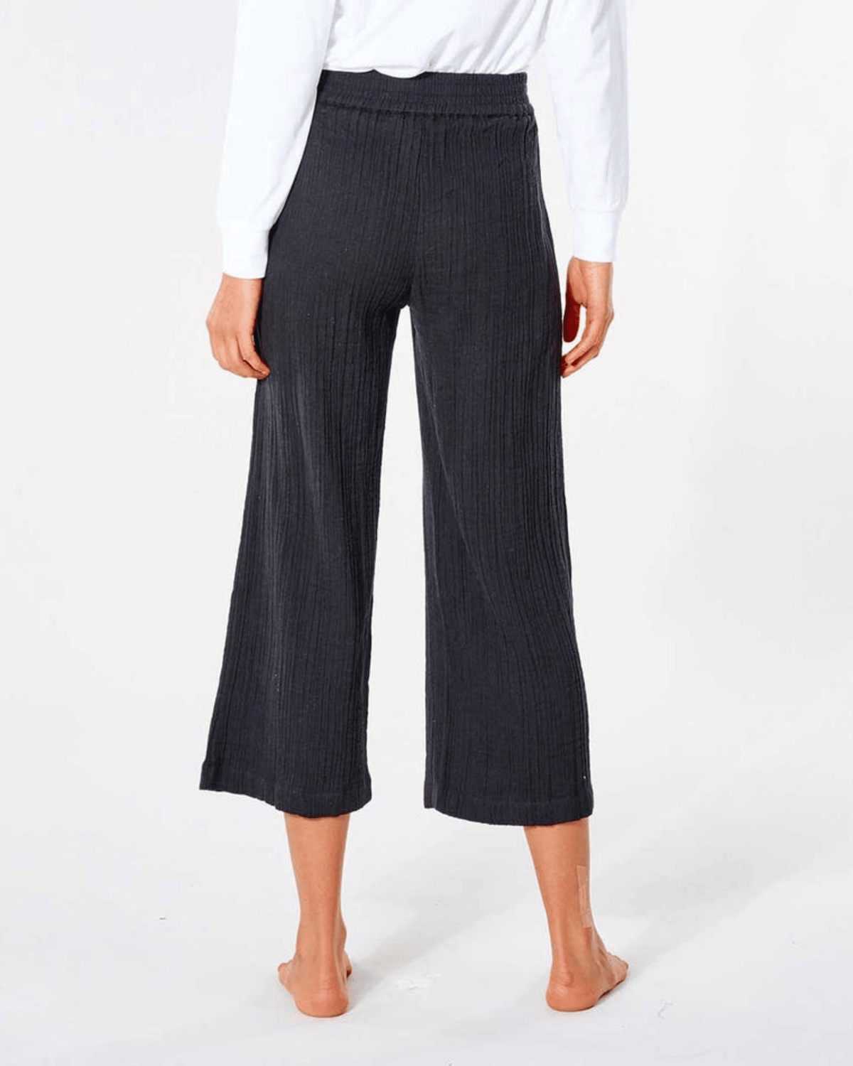 Facing away on a white background, a person is barefoot in black Rip Curl Premium Surf Beach Pants, featuring wide legs and a smocked waist, paired with a white long-sleeve shirt. The cropped pants are ribbed and end at the ankles.
