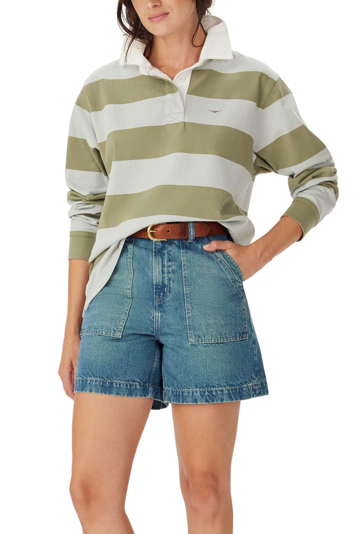 A person wears the RM Williams Narran Stripe Rugby, a loose, long-sleeve green and white striped shirt in heavyweight brushed cotton, tucked into high-waisted denim shorts with large front pockets and a brown belt, hand in pocket.