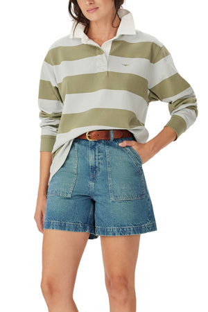 A person wears the RM Williams Narran Stripe Rugby, a loose, long-sleeve green and white striped shirt in heavyweight brushed cotton, tucked into high-waisted denim shorts with large front pockets and a brown belt, hand in pocket.