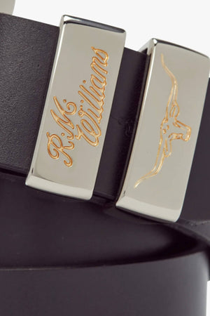 A close-up image of the RM Williams 1 1/2 Jerrawa Belt, made in black solid hide leather in Australia, featuring two polished nickel belt loops. One loop has "R.M. Williams" engraved in gold, and the other features a gold engraved longhorn bull symbol. Its sleek design is completed with a polished nickel buckle.