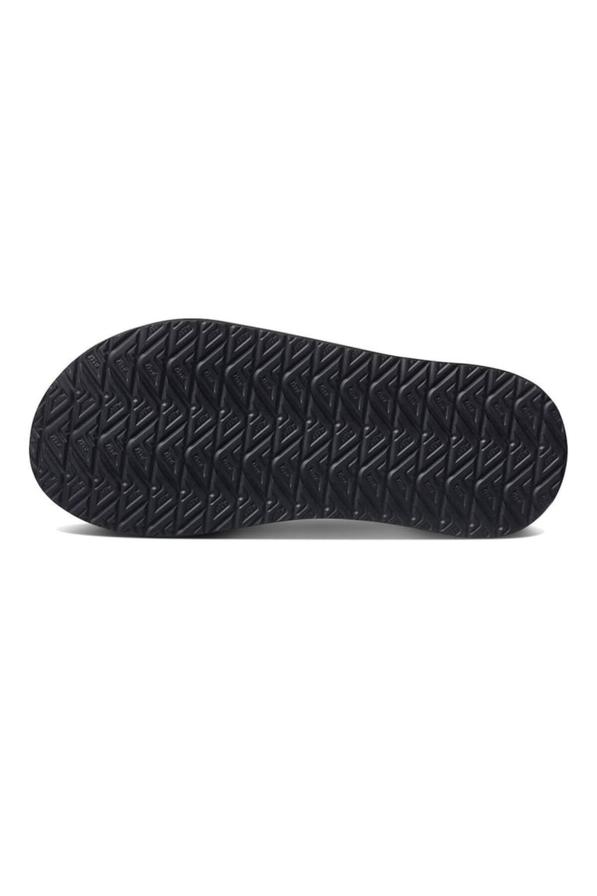 The image displays the bottom of a black REEF Cushion Phantom shoe with a zigzag tread pattern for traction, enhancing durability. Its rugged texture indicates suitability for outdoor or all-weather use, offering comfort with every step.