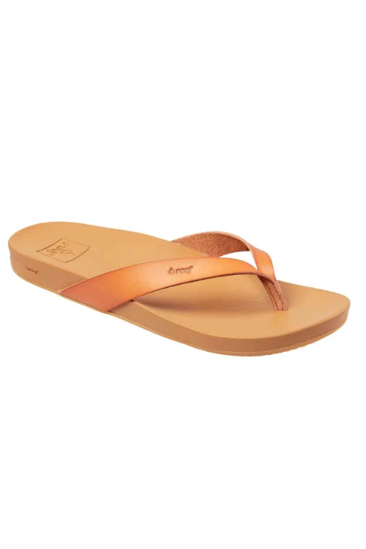 The REEF Cushion Court is a tan flip flop with a flat sole, shiny bronze metallic straps, and gentle arch support, displayed angled on a white background.