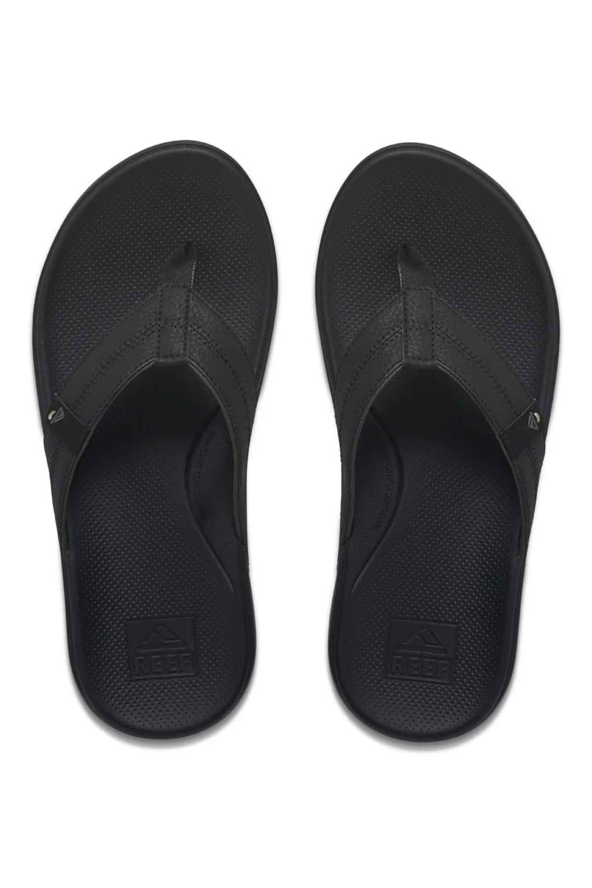 A pair of black REEF Cushion Phantom 2.0 flip-flops with textured footbeds and wide straps, shown from above on a white background. The REEF logo appears on the heel, and these sandals are designed for all-day comfort.