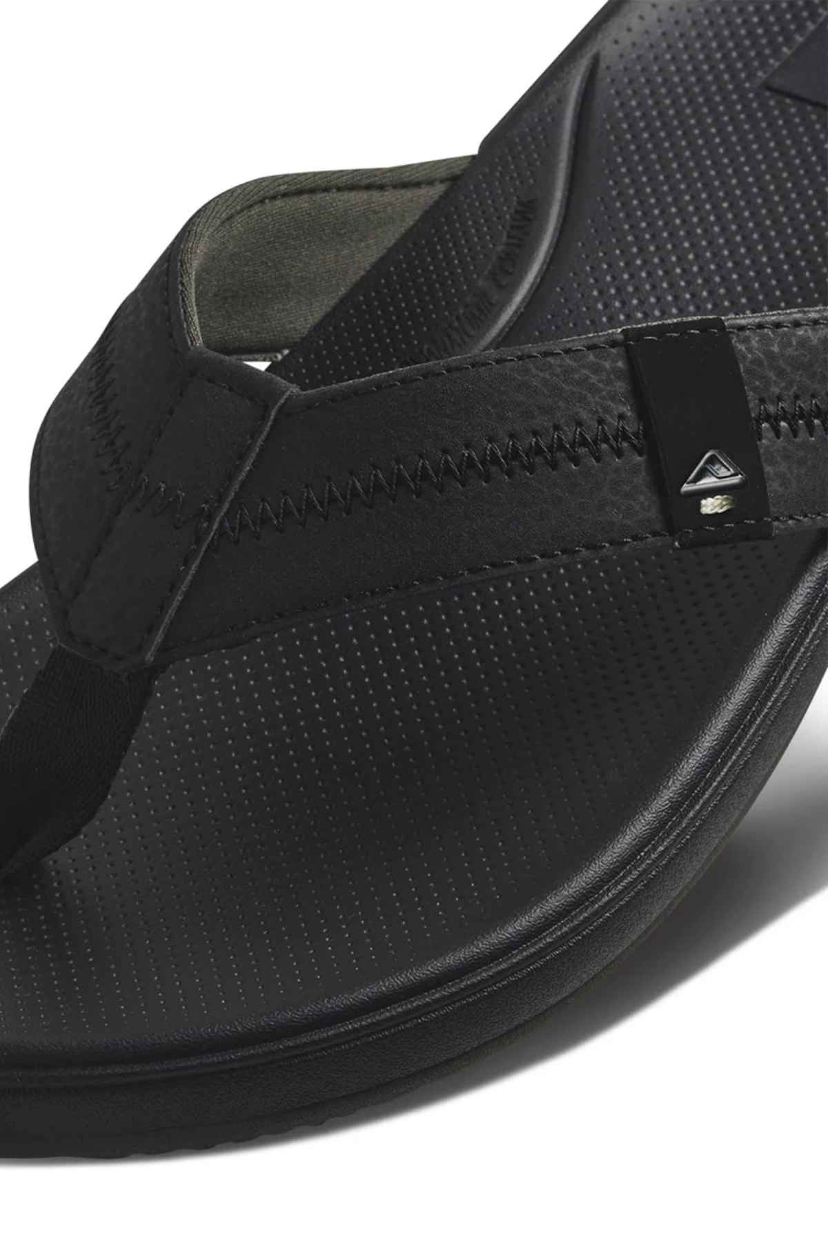 Close-up of a black REEF Cushion Phantom 2.0 flip-flop featuring a textured footbed, visible strap stitching, arch support, and a small triangular metallic logo detail on the side.