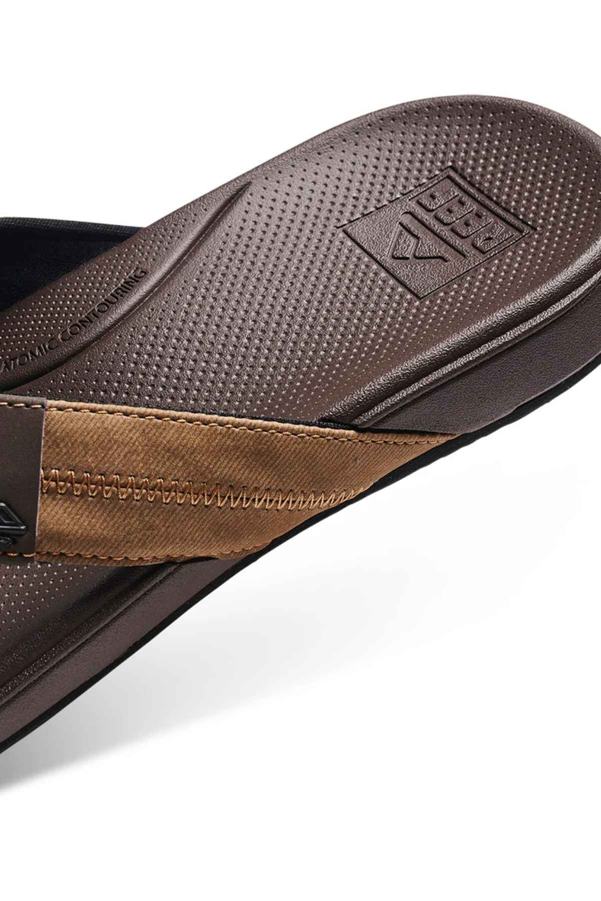 Close-up of the brown REEF Cushion Phantom 2.0 sandal featuring a textured footbed, tan fabric strap, molded rubber outsole, and embossed brand logo near the heel.