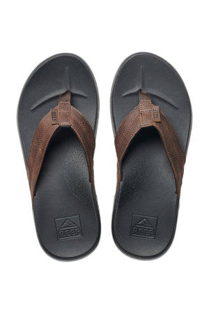 The REEF Cushion Phantom LE sandals feature wide brown leather straps and a black thong design, shown side by side on a white background. The cushion footbed has textured surfaces and displays the "REEF" logo.