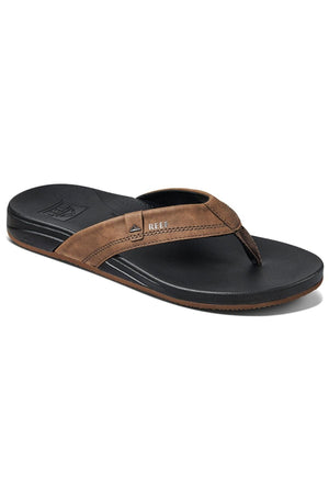 The REEF Cushion Spring is a brown and black thong sandal with a textured footbed providing integrated arch support. Its synthetic leather strap has a small label, perfectly matching the black sole's subtle tread. Angled for style, it's both comfortable and fashionable.