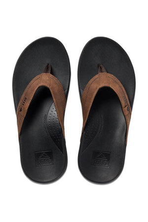 Top view of a pair of REEF Cushion Spring thongs. The brown synthetic leather straps with the brand logo contrast the black soles, which feature textured insoles for grip and arch support.