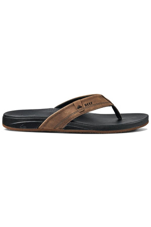 The REEF Cushion Spring is a brown and black flip-flop sandal with synthetic leather straps featuring visible stitching. It has a black sole that provides slight arch support and an elevated heel, while the brand logo decorates the strap. Ideal for simple, casual everyday wear.