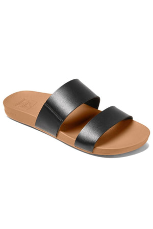 The REEF Cushion Vista is a stylish tan sandal featuring two wide, black leather straps that cross over the foot. It has a flat sole crafted from smooth, cushioned material with an EVA foam compound for extra comfort. This minimalist design also includes an open toe.