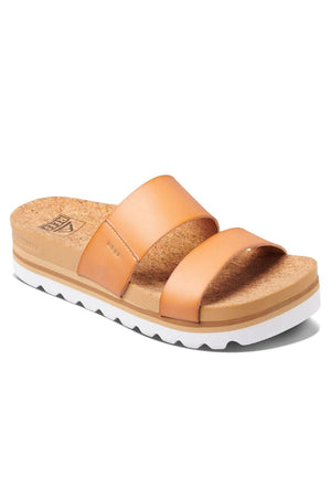 The REEF Cushion Vista Hi sandal showcases two wide leather straps over a cork footbed and features a white and tan zigzag-patterned sole. It offers all-day comfort with its cushioned platform heel, while branding is visible on both the strap and the side of the sole from the left view.