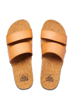 A pair of tan REEF Cushion Vista Hi slip-on sandals with two wide straps across the top, featuring cork insoles and a subtle platform heel. The brand name "REEF" and its logo are visible on the cushioned insole.