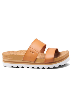 The REEF Cushion Vista Hi is a tan-colored sandal featuring a cushioned cork footbed and two broad straps across the top. The sole is layered with a white, slightly wavy grip on the bottom, making it perfect for casual wear.