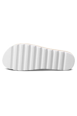 The image displays the underside of the REEF Cushion Vista Hi, showcasing a white sole. This sole features a ripple design with parallel grooves running across it, offering texture and presumably good traction. The clean, cushioned sandal's sole also has a slight curve to its shape.