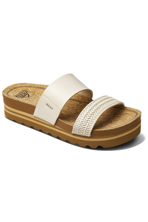 The REEF Cushion Vista Hi TX is a brown sandal with a cork-like textured insole and a thick, ridged sole. It features two off-white straps: the front one is braided, and the larger one closer to the ankle is smooth, with the brand name "REEF" visible on it. Enjoy sustainable footwear with added comfort from its cushion midsole.