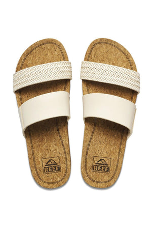 A pair of beige REEF Cushion Vista Hi TX slip-on sandals with cork footbeds and two wide straps across each foot. The upper strap features a woven pattern, while the lower strap is solid. The cushion midsole ensures added comfort, and the "Reef" brand logo is printed on the footbeds near the heel area.