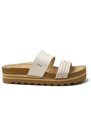 The REEF Cushion Vista Hi TX is a sustainable tan platform sandal featuring a cork footbed and black ridged sole. It includes two white straps: one wide strap near the toes adorned with a chevron pattern, and a plain, narrower strap nearer to the middle embossed with "REEF Cushion Vista." The sandal also boasts a cushion midsole for enhanced comfort.