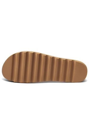 The image displays the underside of a brown rubber shoe sole from the REEF Cushion Vista Hi TX, characterized by its wavy, ribbed texture running horizontally. Deep grooves and ridges define the pattern, enhancing traction. Part of REEF's sustainable footwear line, this shoe includes a cushioned midsole for extra comfort.