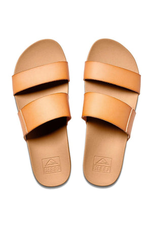 A top-down view of the REEF Cushion Vista sandals featuring tan, vegan leather straps and a minimalist design. The open-toe sandals showcase "REEF" branding on the footbeds. With their cushion bounce midsole and flat sole, they offer style with no visible arch support.