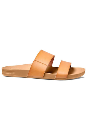 The REEF Cushion Vista is a tan leather slide sandal with two vegan leather straps across the top and a flat sole. It boasts a simple and minimalist design that is perfect for casual wear, complemented by a cushion bounce midsole for enhanced comfort.