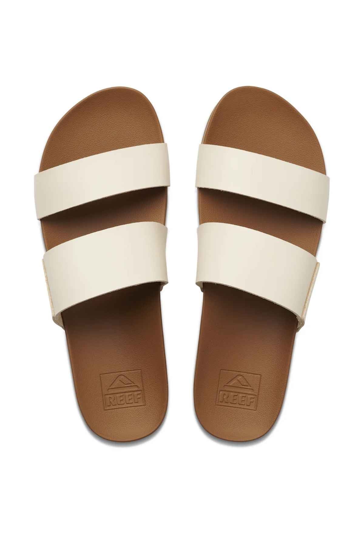 A pair of REEF Cushion Vista brown slide sandals with minimalist design features two wide cream straps, shown from above. The footbed offers memory-free foam and arch support, with the brand name embossed on it.
