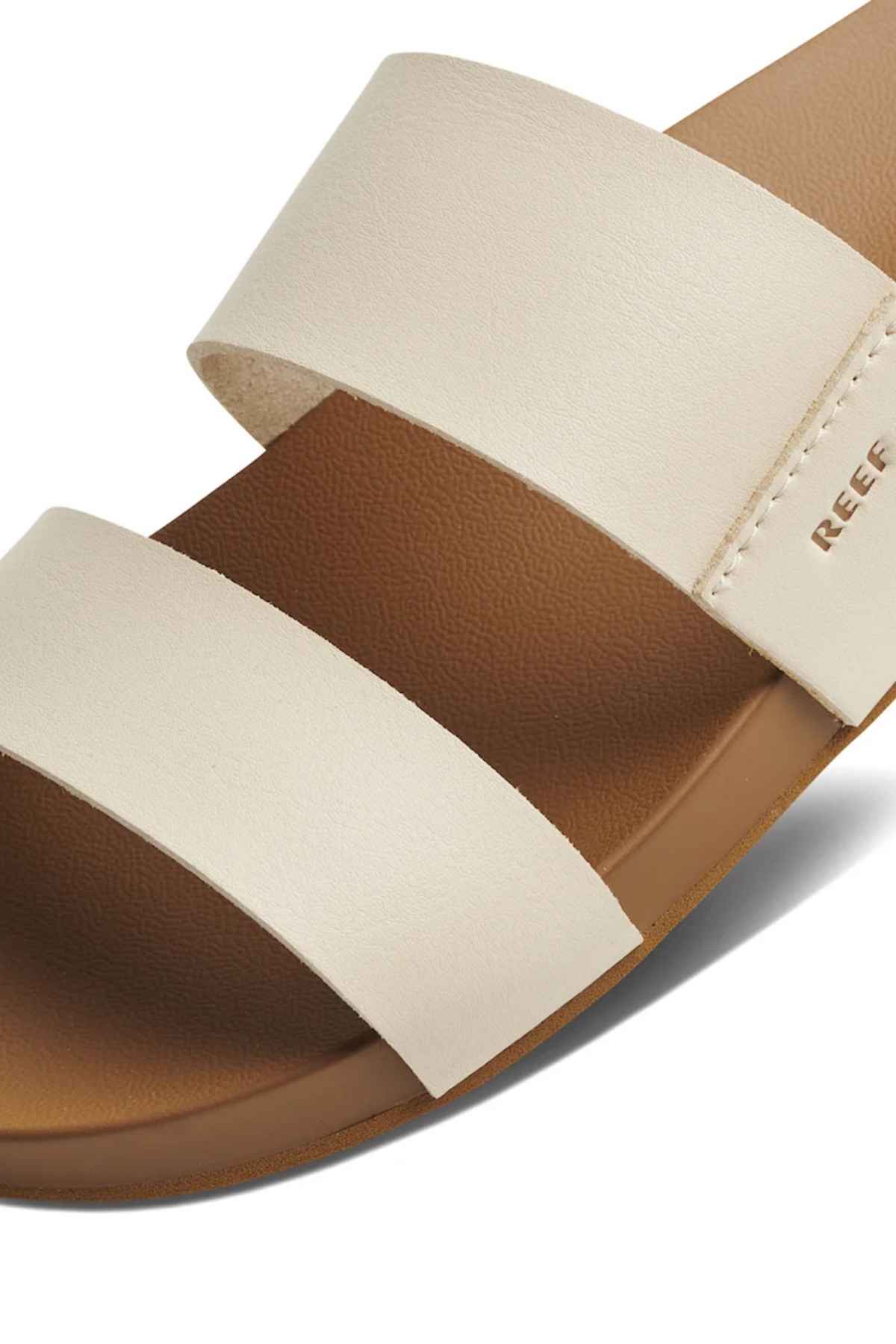 The REEF Cushion Vista sandal has a close-up view showing two wide beige straps, a brown sole, memory-free foam footbed for comfort, visible arch support, and the "REEF" brand name on the side strap.