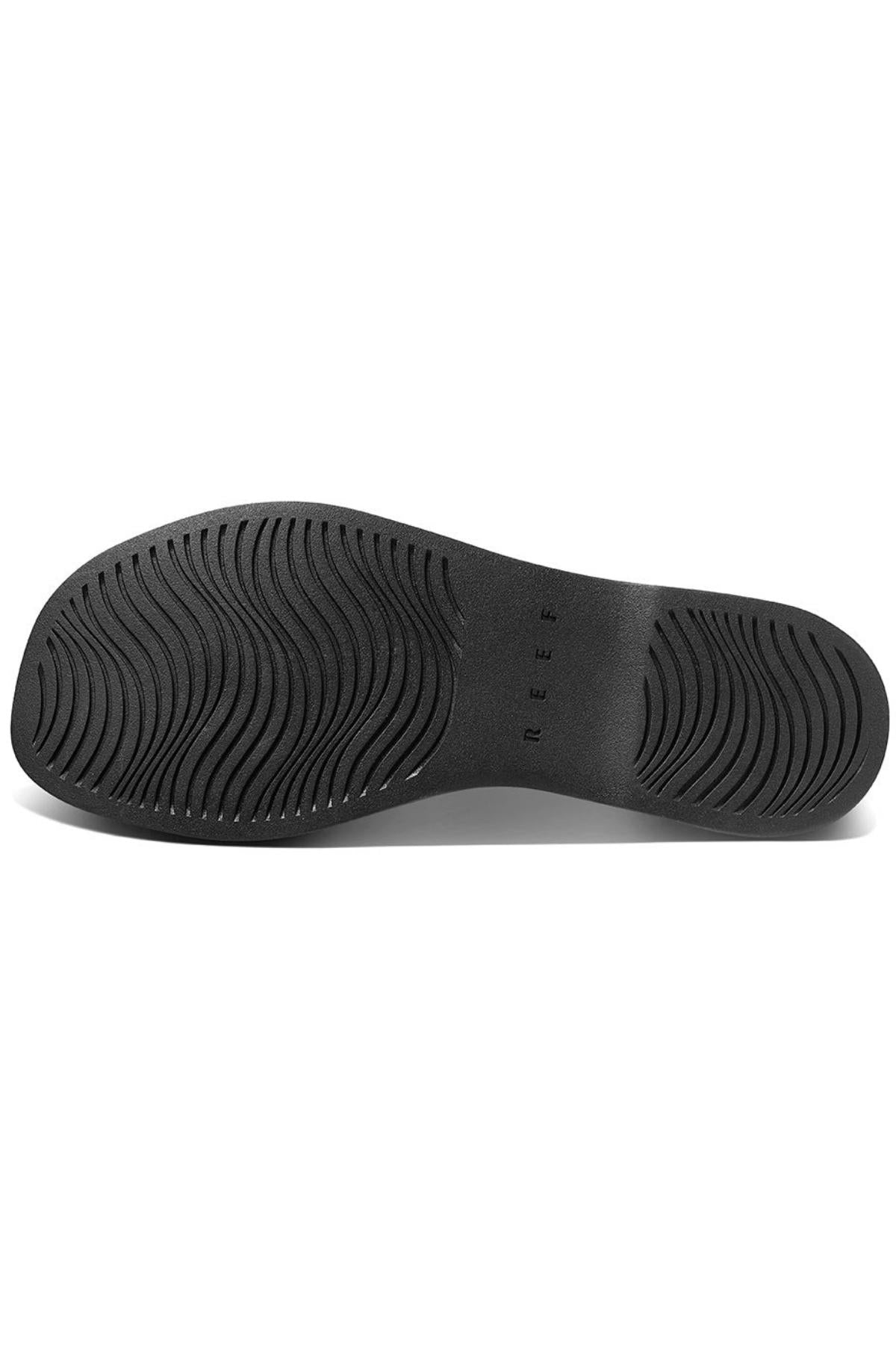 The image shows the black rubber sole of the REEF Lofty Lux X shoe, with wavy textured patterns for grip and "REEF" embossed near the center, highlighting its sustainable design and premium comfort.