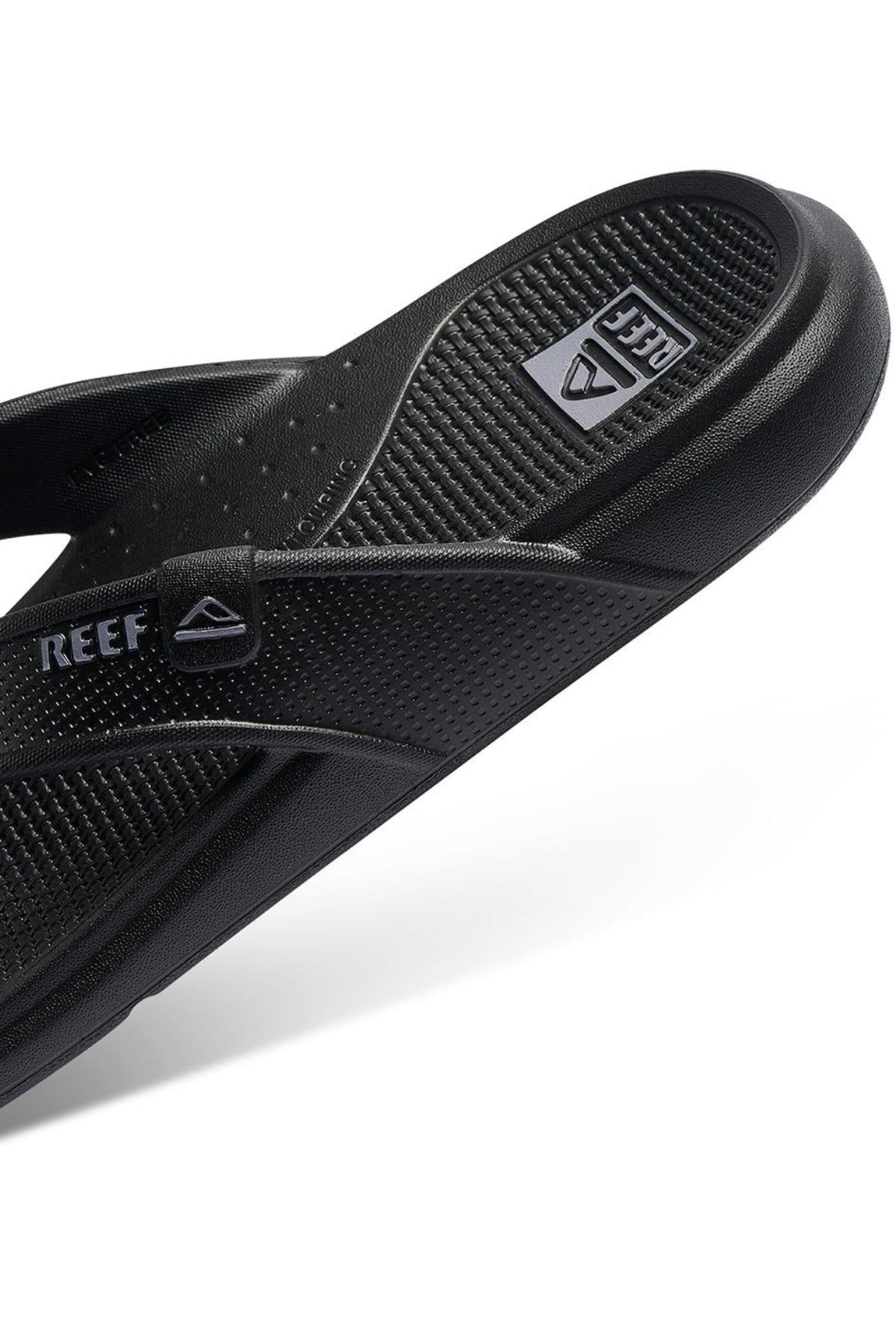 A close-up view of the black REEF Oasis sandal highlights its textured footbed and strap. The white letters of the Reef logo are clearly visible on the strap, while the brand's name is subtly etched into the footbed. Made with renewable sugarcane EVA and offering arch support, this sandal boasts a minimalist yet sporty design.