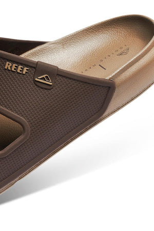 Close-up of the REEF Oasis Double Up sandal in brown, featuring textured material, brand logo, and a renewable sugarcane EVA footbed with arch support on a white background.