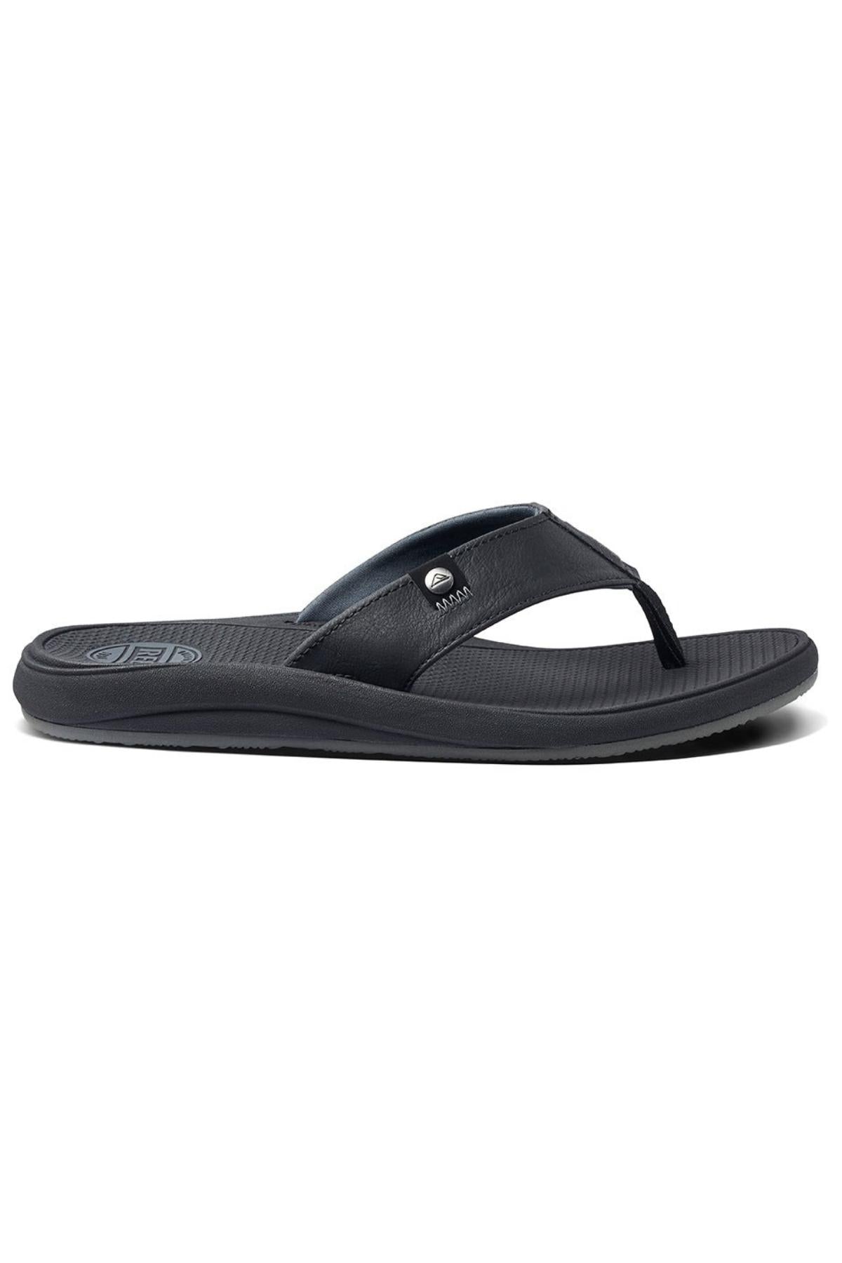 A side view of the REEF Phantom Nias flip-flop sandal showcases its sleek black design, featuring a textured footbed and a minimalist strap. The strap is adorned with a small metal logo embellishment. It also includes a slightly raised heel for enhanced support and a durable outsole that promises a comfortable fit.