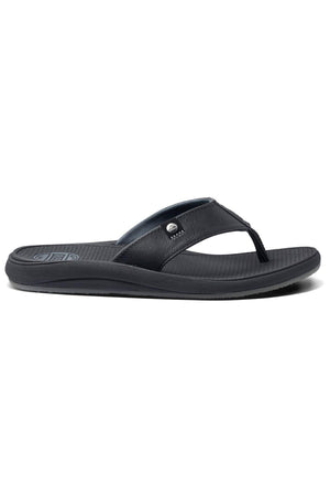 A side view of the REEF Phantom Nias flip-flop sandal showcases its sleek black design, featuring a textured footbed and a minimalist strap. The strap is adorned with a small metal logo embellishment. It also includes a slightly raised heel for enhanced support and a durable outsole that promises a comfortable fit.