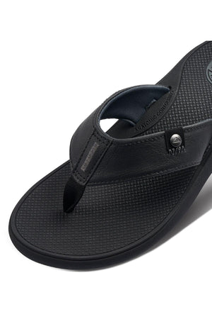 A close-up of the REEF Phantom Nias flip flop sandal in black, showcasing its textured footbed and robust thong strap. The sandal is embellished with metal rivets by the strap and features discreet branding on both the footbed and strap. Designed with longevity in mind, it promises a comfortable fit and durable outsole.