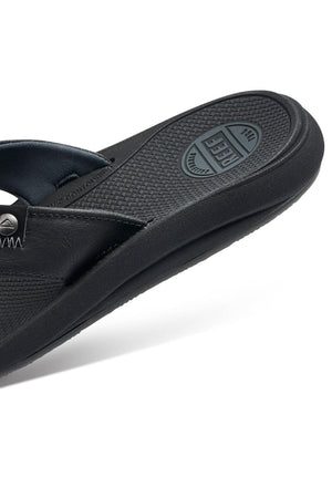 A close-up view of the REEF Phantom Nias sandal in black, showcasing its textured sole with visible branding. The sandal features a robust construction with detailed stitching along the strap and a durable outsole for added reinforcement. Photographed against a plain white background, the design and features of the sandal are highlighted.